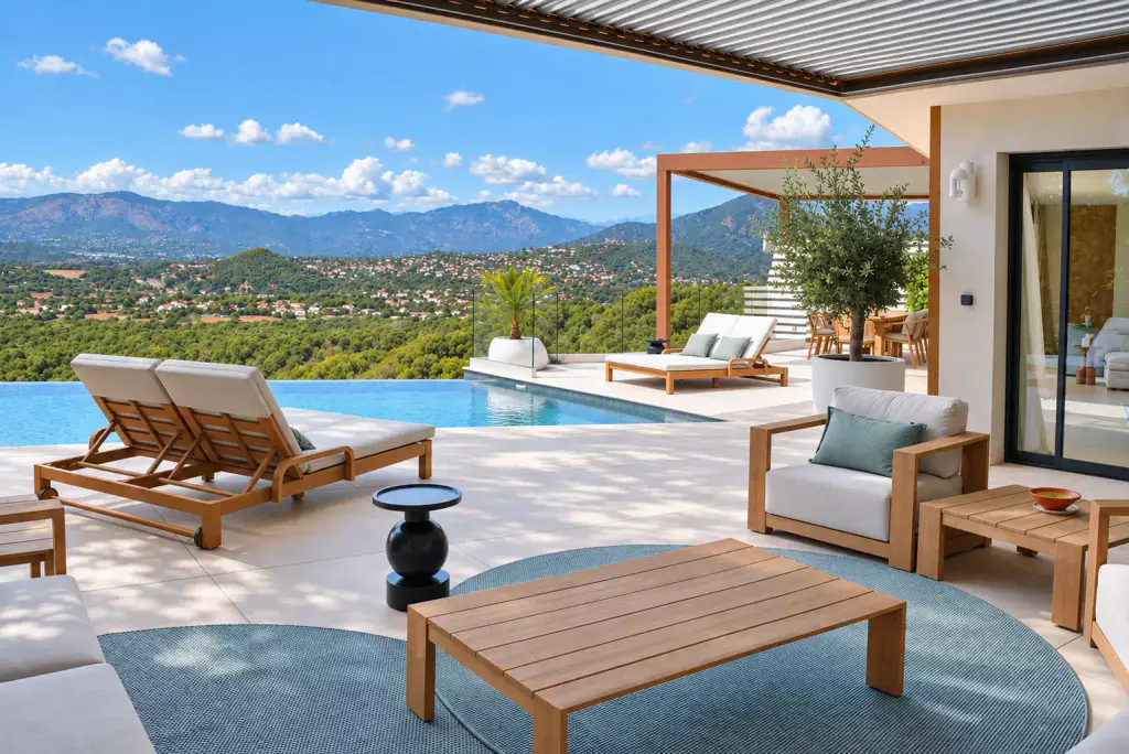 Salon d’extérieur en teck sur un tapis rond bleu-vert, sous la pergola : bains de soleil au bord de la piscine à débordement, seconde pergola et montagnes au fond.