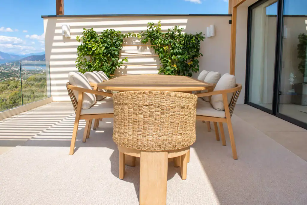Teak dining table surrounded by rope chairs and a woven armchair, under the pergola; against the white wall striped with shadows, two espaliered citrus trees in planters.