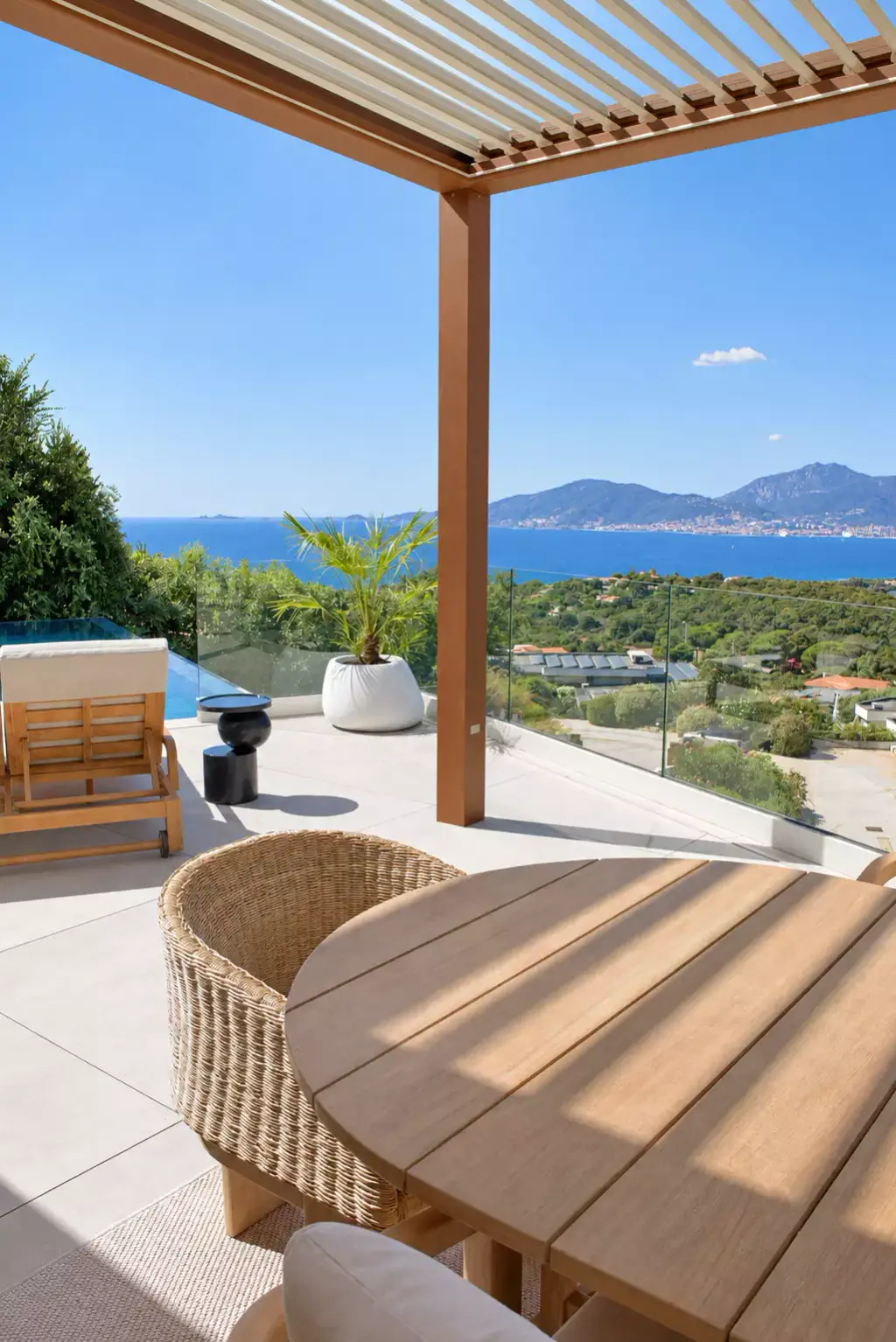 Table ronde en teck et fauteuil en fibre tressée sous la pergola aux lames entrouvertes ; entre deux poteaux brun cuivré, le garde-corps vitré, puis le golfe.