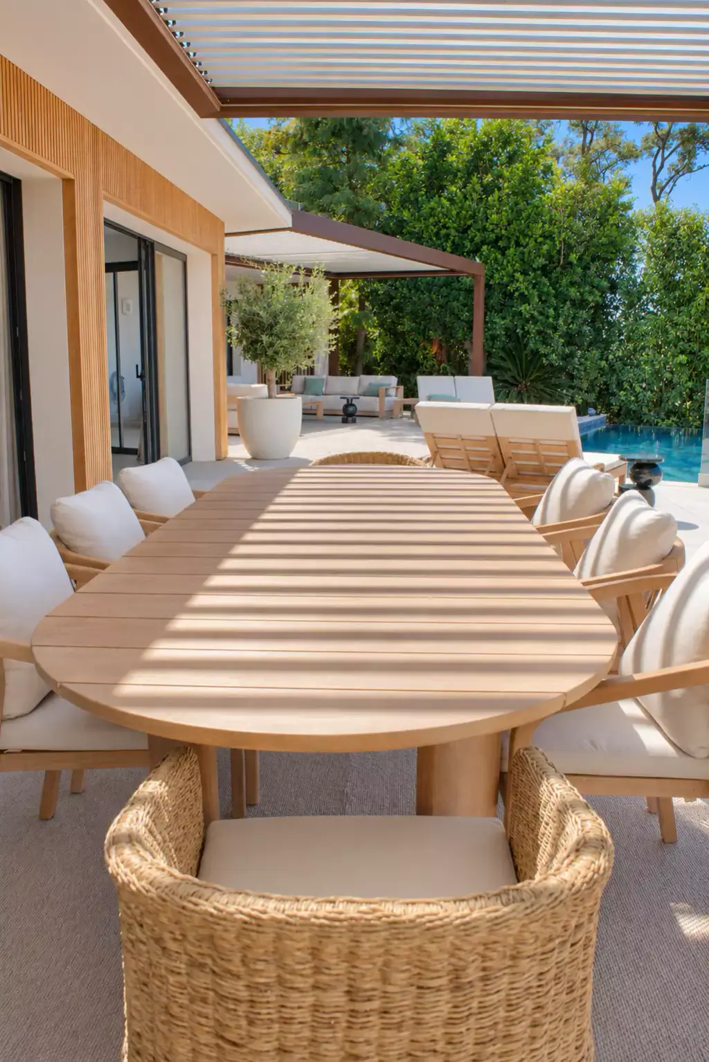 Grande table ovale en teck rayée par l’ombre des lames, fauteuil en fibre tressée au premier plan ; la terrasse file vers la seconde pergola et la piscine.