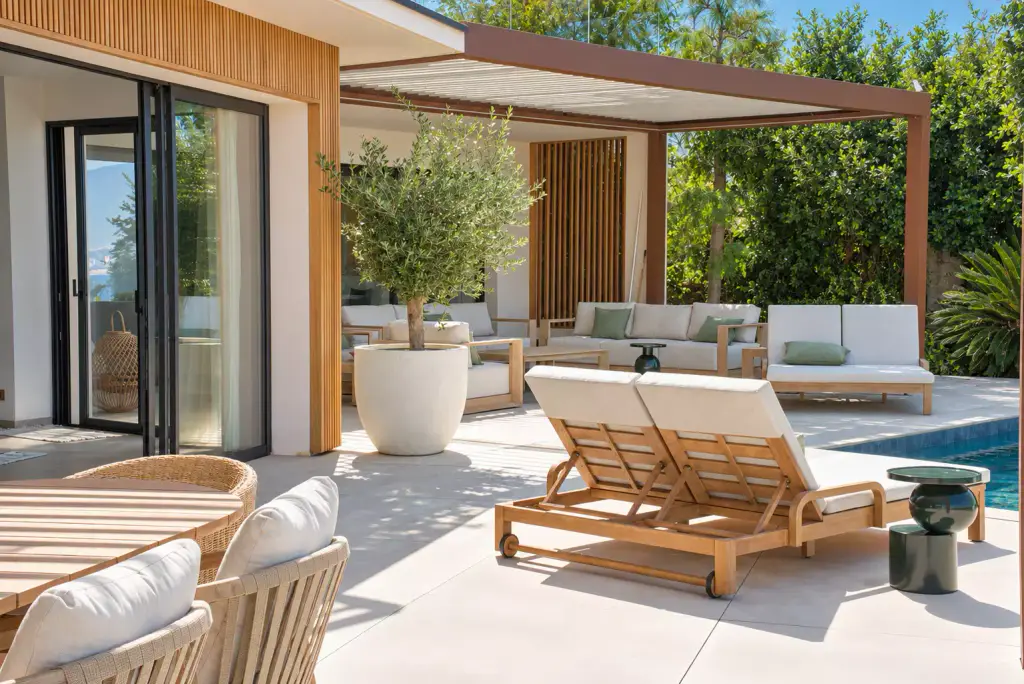 Terrasse le long de la façade habillée de tasseaux de bois : chaises et bain de soleil double en teck, olivier en pot, salon d’extérieur sous la pergola brun cuivré.