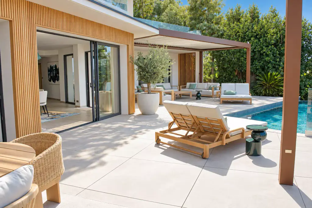 Terrace between house and pool: open glass door in the wooden-batten façade, double teak sun lounger, potted olive tree and lounge under the pergola.