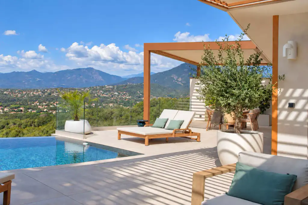 Terrasse de piscine à débordement face aux montagnes : bain de soleil double en teck, olivier en pot blanc et pergola bioclimatique brun cuivré au fond.