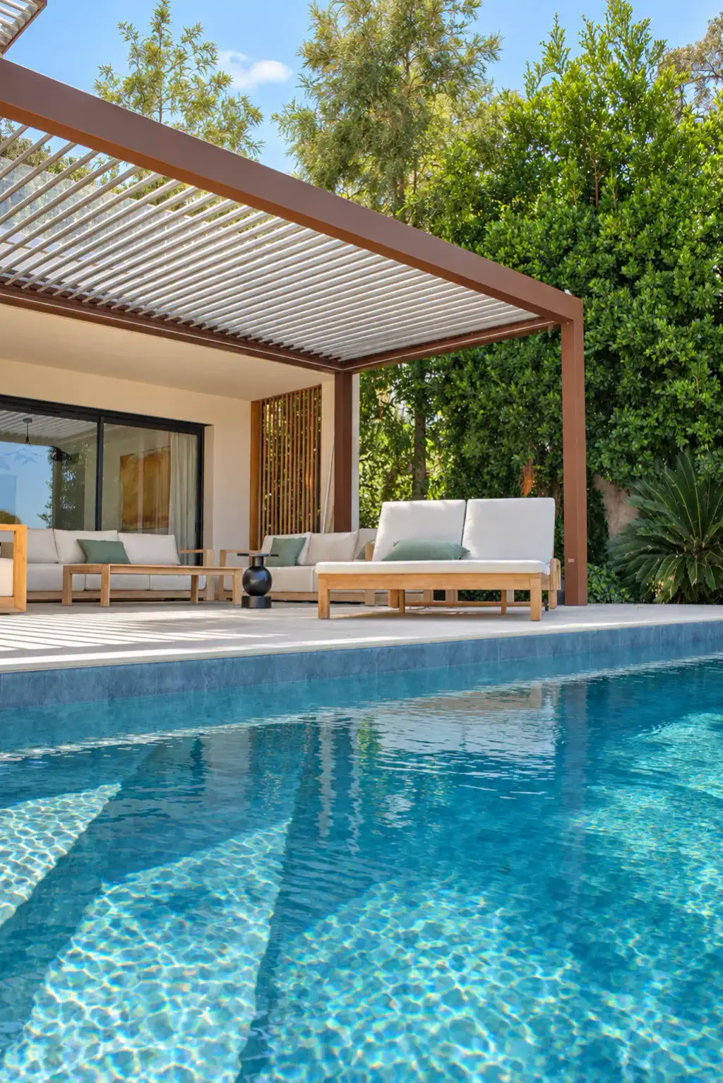 Piscine au bleu clair au premier plan ; sur la terrasse, le salon d’extérieur en teck à l’abri de la pergola bioclimatique brun cuivré, contre une haie dense.