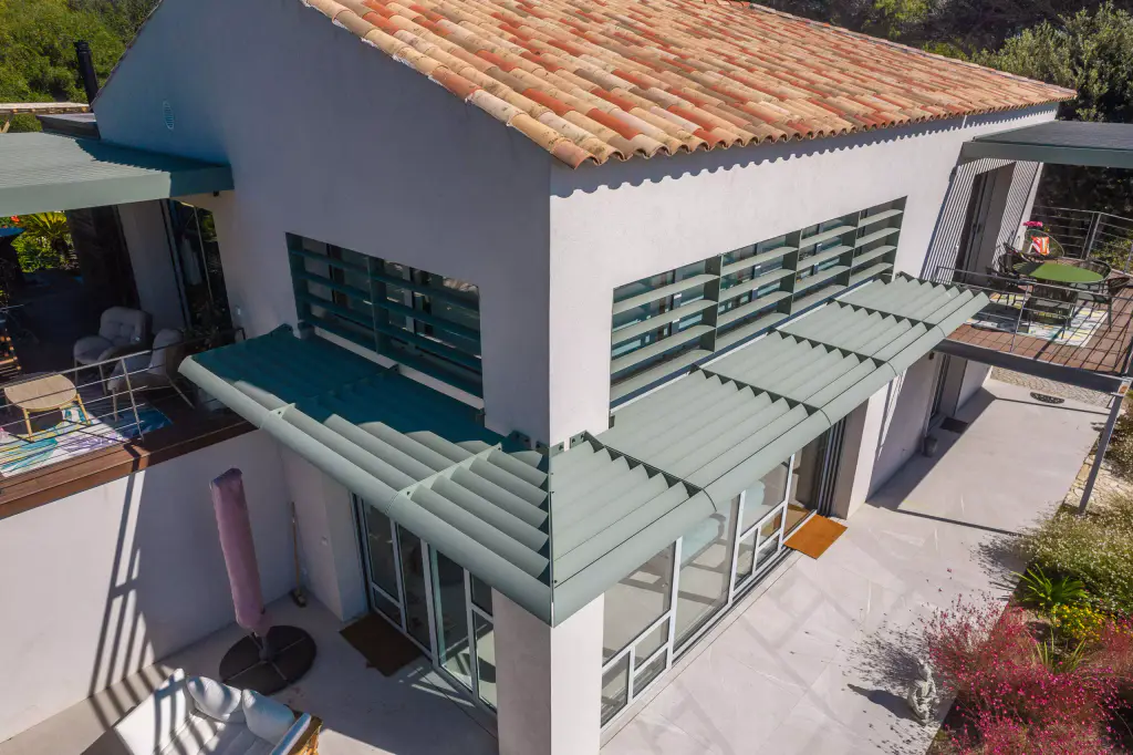 Casquette brise-soleil à lames vert sauge qui tourne l’angle au-dessus de la verrière, lames vertes devant les fenêtres hautes, mur blanc et toit de tuiles ; terrasse dallée en bas.