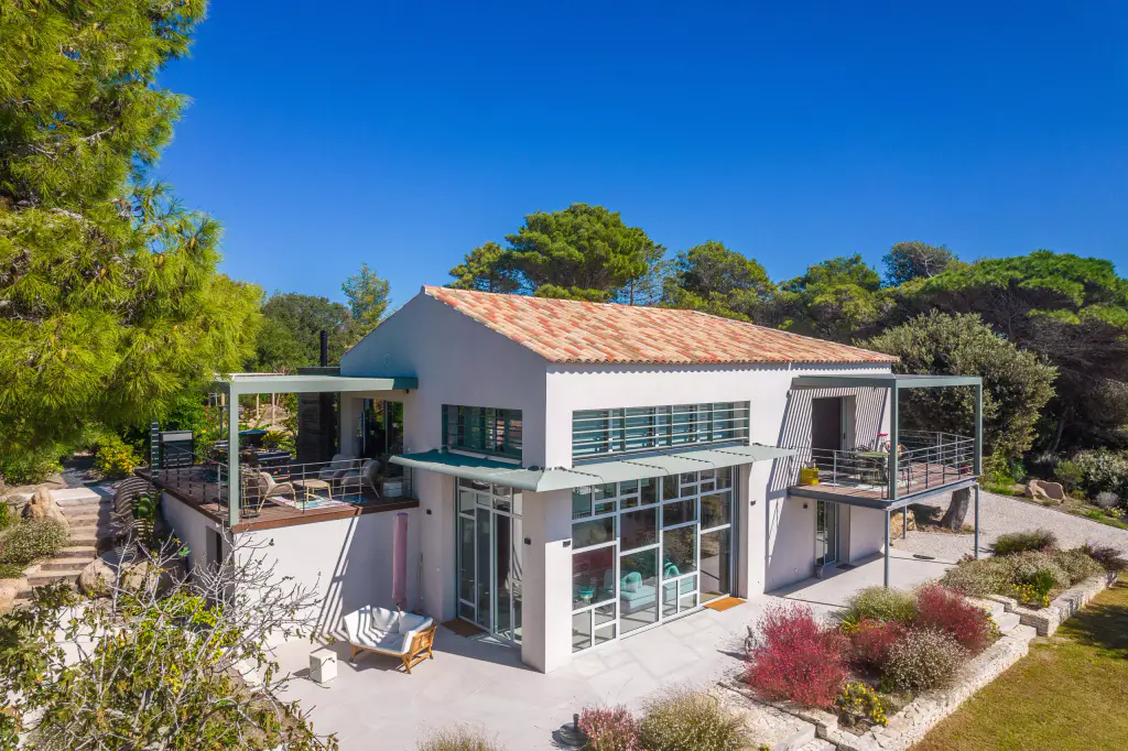 Vue drone de trois quarts : maison blanche à toit de tuiles, pergola verte sur chaque terrasse d’étage, casquette à lames vertes au-dessus de la verrière, massifs fleuris.