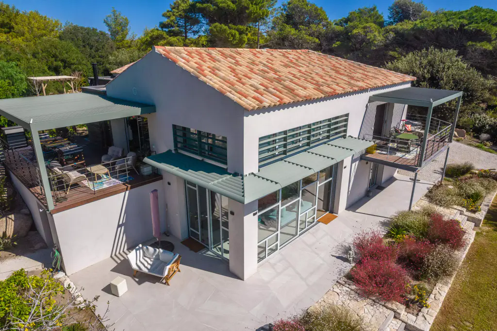 Vue drone de trois quarts : pergola verte sur la terrasse en bois à gauche, casquette brise-soleil à lames vertes sur la verrière, seconde pergola à droite, toit de tuiles.