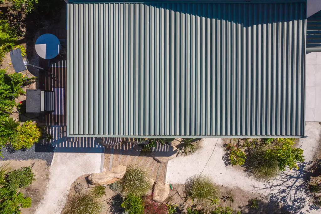 Vue drone à la verticale : toiture vert sauge de la pergola, lames fermées, qui remplit le haut de l’image ; brasero bleu et deck à gauche, allée claire et rocaille en bas.