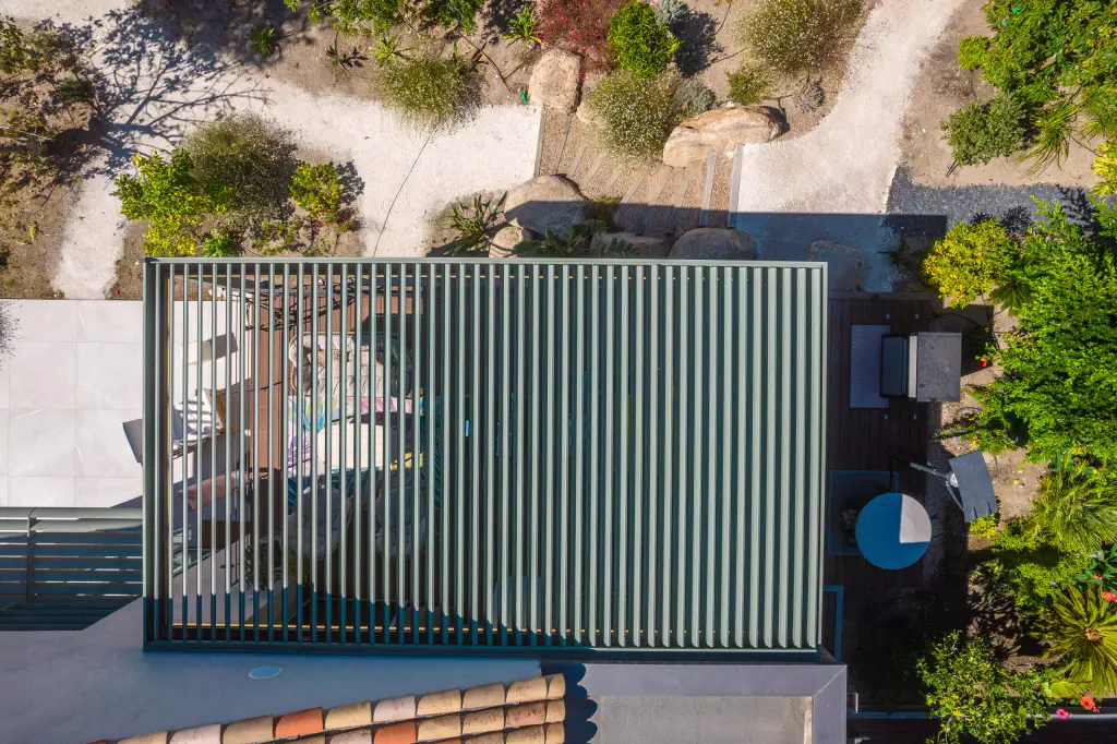 Vue drone à la verticale : lames vert sauge de la pergola ouvertes, le salon et son tapis coloré se lisent au travers, rochers et rocaille autour.