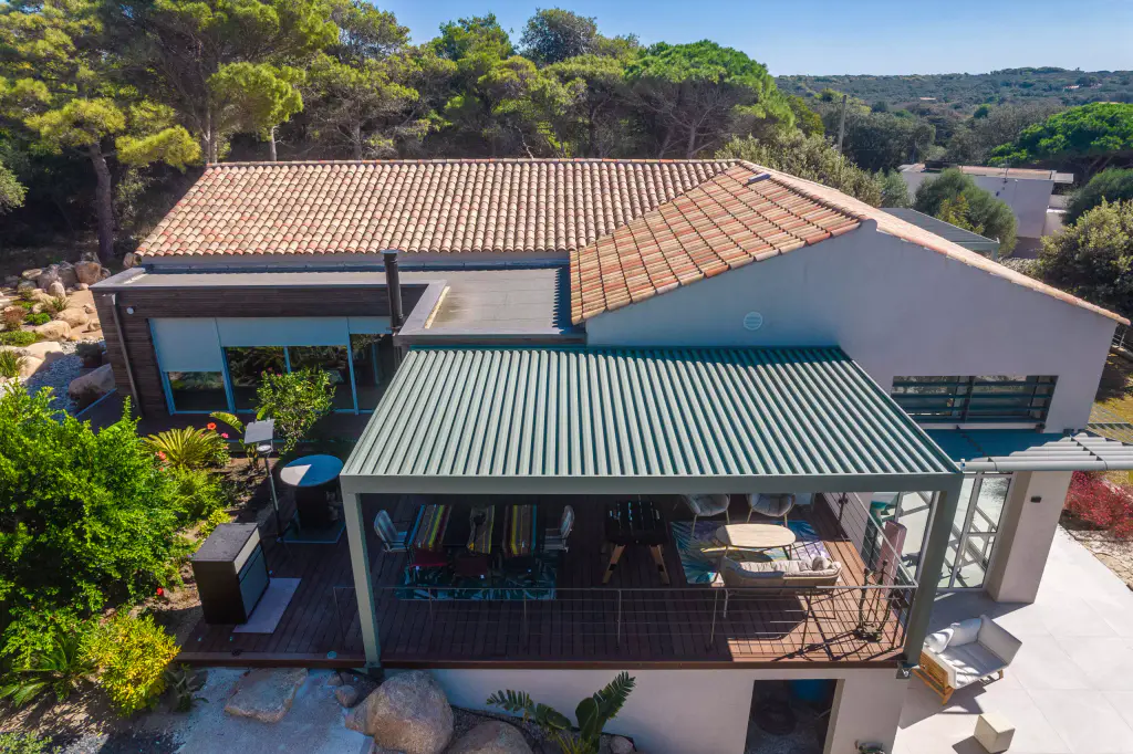 Vue drone plongeante : pergola vert sauge aux lames fermées adossée à la maison blanche à toit de tuiles, terrasse en bois meublée dessous, garde-corps à câbles, pins au fond.