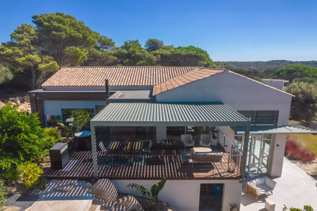 Vue drone de face : pergola vert sauge, lames fermées, sur la terrasse en bois de la maison blanche ; table à manger, fauteuils clairs et canapé dessous, toit de tuiles et pins.