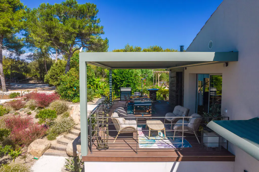 Pergola bioclimatique vert sauge sur une terrasse en bois d’étage : fauteuils et canapé en corde claire, tapis coloré, table à manger au fond, escalier de pierre et massifs fleuris à gauche.