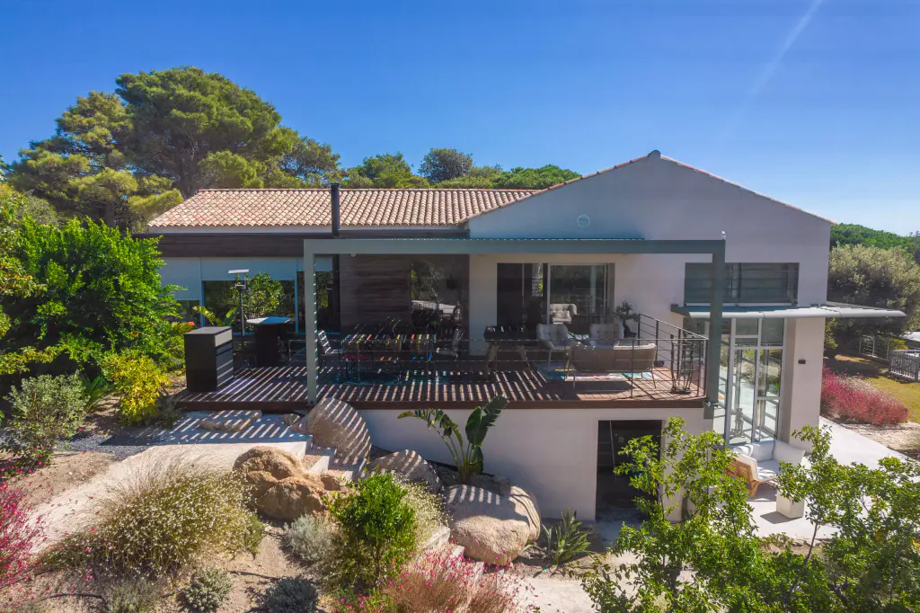 Maison blanche à toit de tuiles et pergola vert sauge sur la terrasse en bois d’étage, vues depuis le jardin : rocaille, graminées et massifs à floraison rouge au premier plan.
