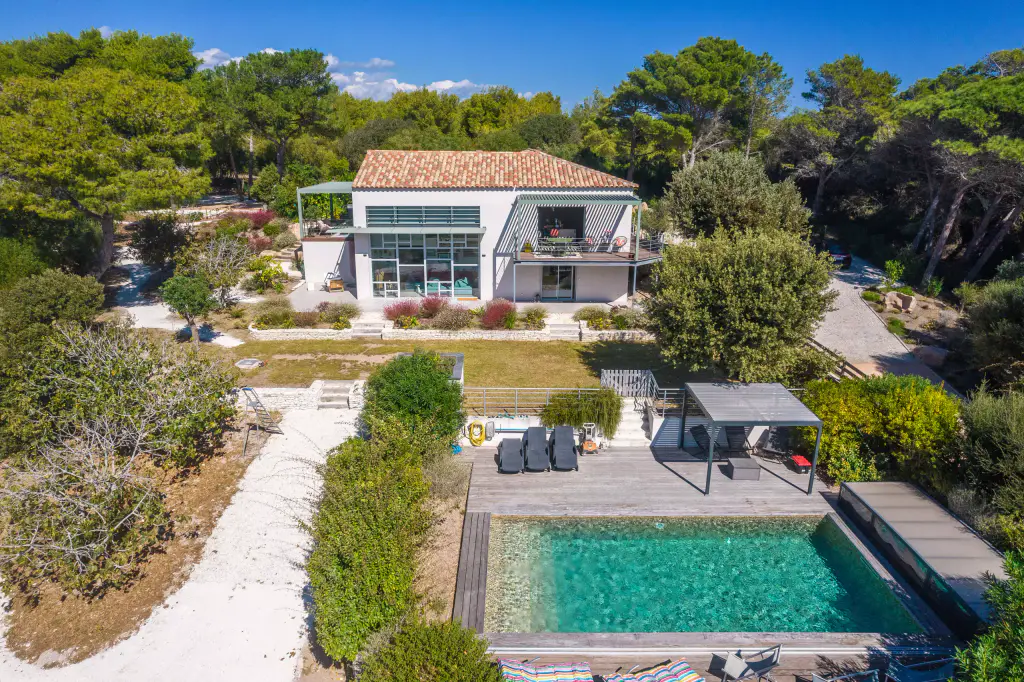 Piscine vert d’eau bordée d’un deck en bois, bains de soleil et petite pergola grise au premier plan ; au fond, la maison blanche à toit de tuiles et ses pergolas vertes, entre les pins.