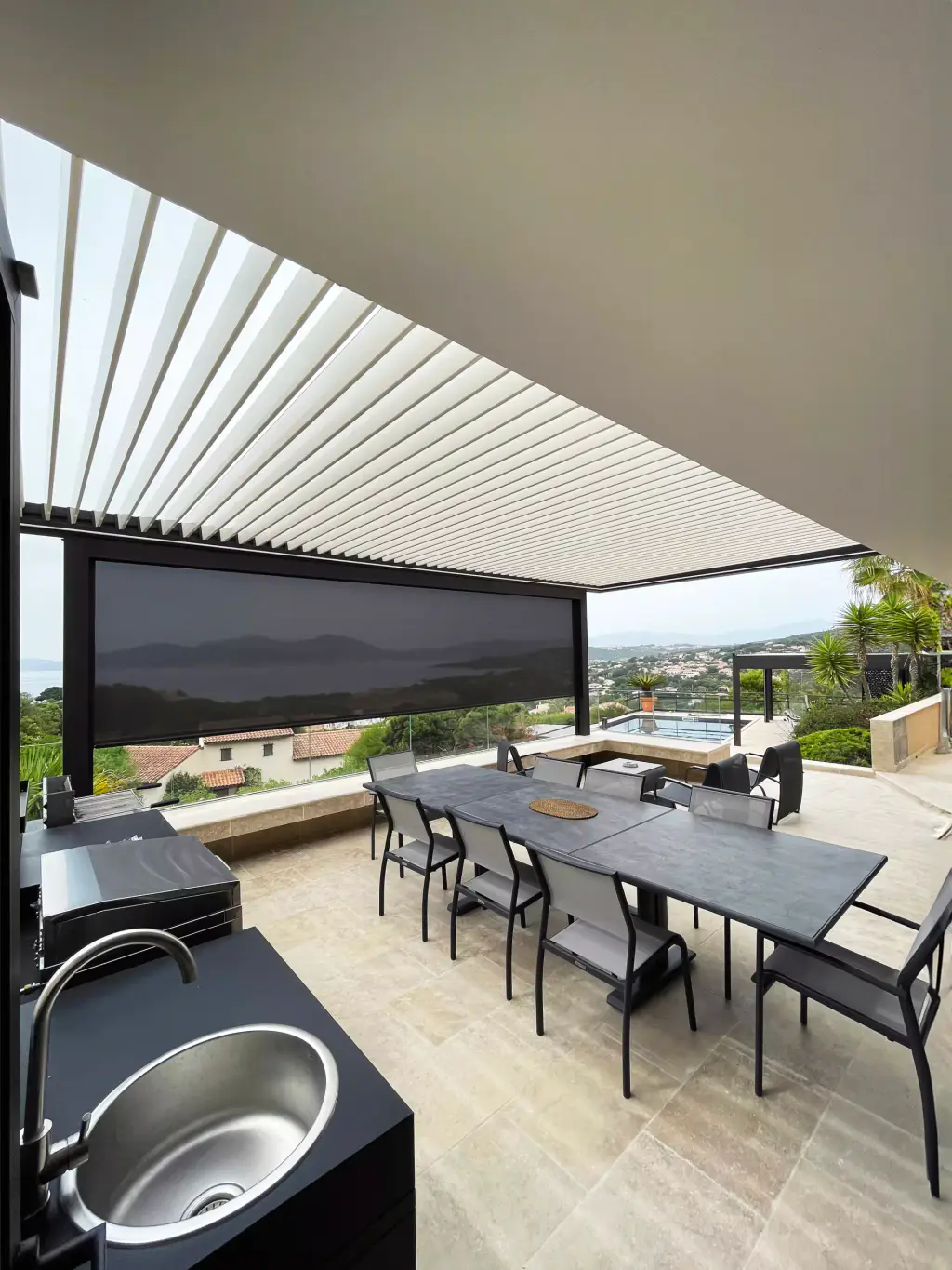 Cuisine d’été sous la pergola : évier inox au premier plan, table à manger, store vertical descendu au travers duquel on devine le golfe.