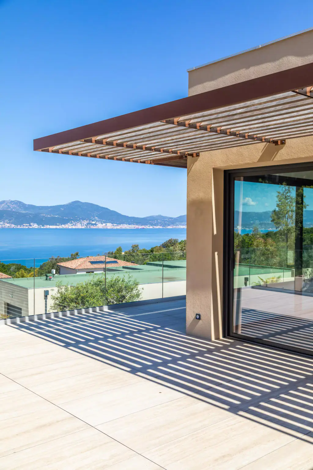 Brise-soleil en casquette à lames de bois, cadré en hauteur : l’angle de la villa enduite ocre, la baie vitrée, les ombres rayées sur la terrasse et le golfe à gauche.