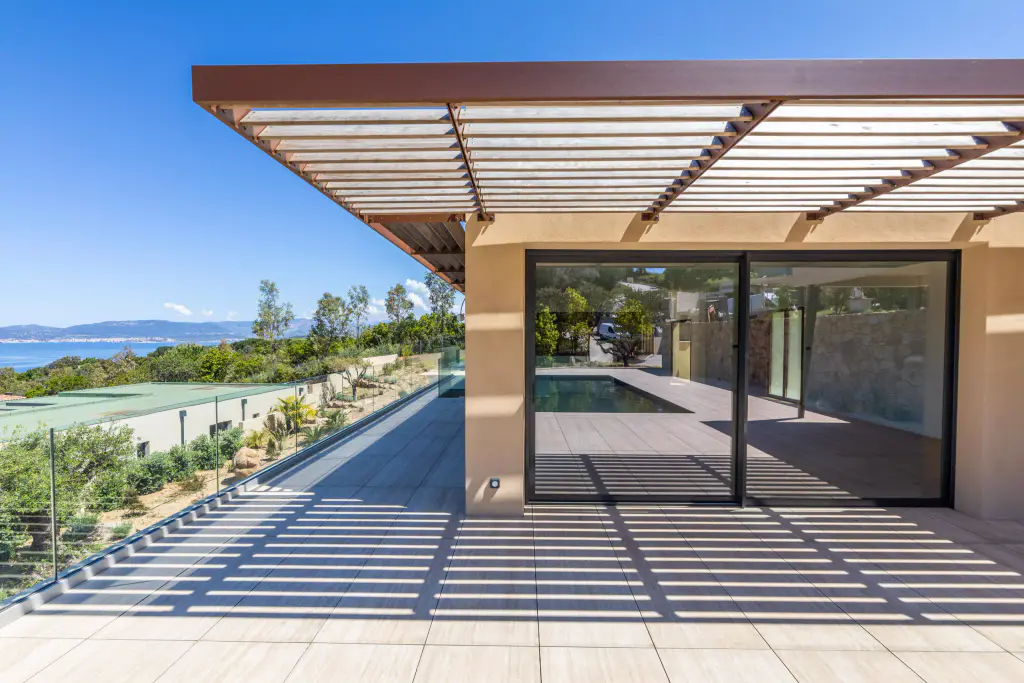 Brise-soleil en casquette à lames de bois et rive d’acier brun au-dessus de la baie vitrée, vu de trois quarts ; coursive carrelée rayée d’ombres, jardin et golfe à gauche.