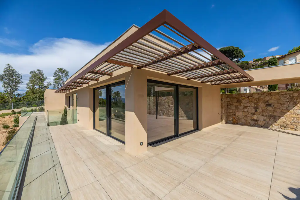 Corner of the ochre flat-roofed villa: the wooden louvred brise-soleil canopy runs along two façades, sliding glass doors, tiled terrace, stone wall on the right.