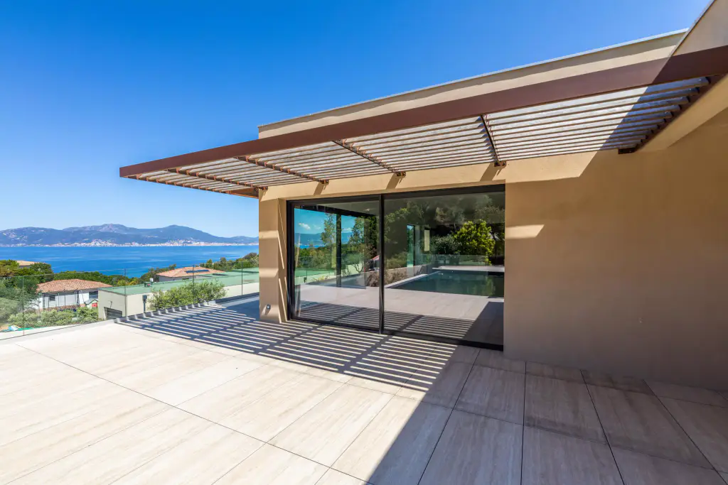 Grande terrasse carrelée claire devant la villa ocre à toit plat : brise-soleil en casquette à lames de bois au-dessus de la baie coulissante, ombres rayées, golfe et montagnes.