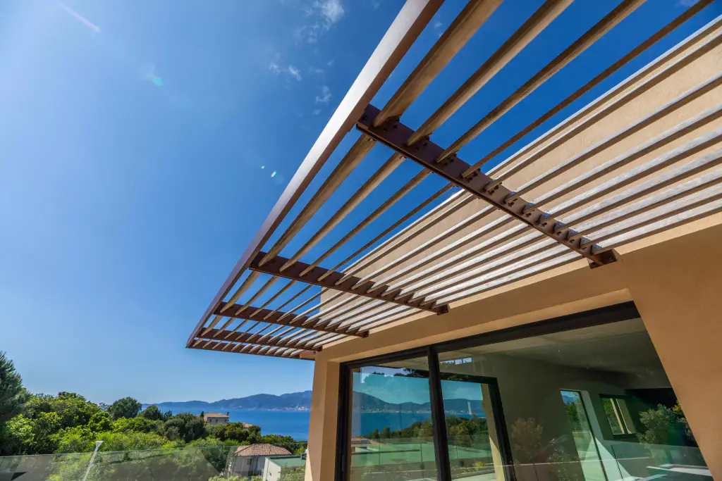 Brise-soleil en contre-plongée : la casquette à lames de bois et consoles d’acier brun s’avance sur un ciel bleu profond au-dessus de la baie vitrée, golfe et montagnes à gauche.