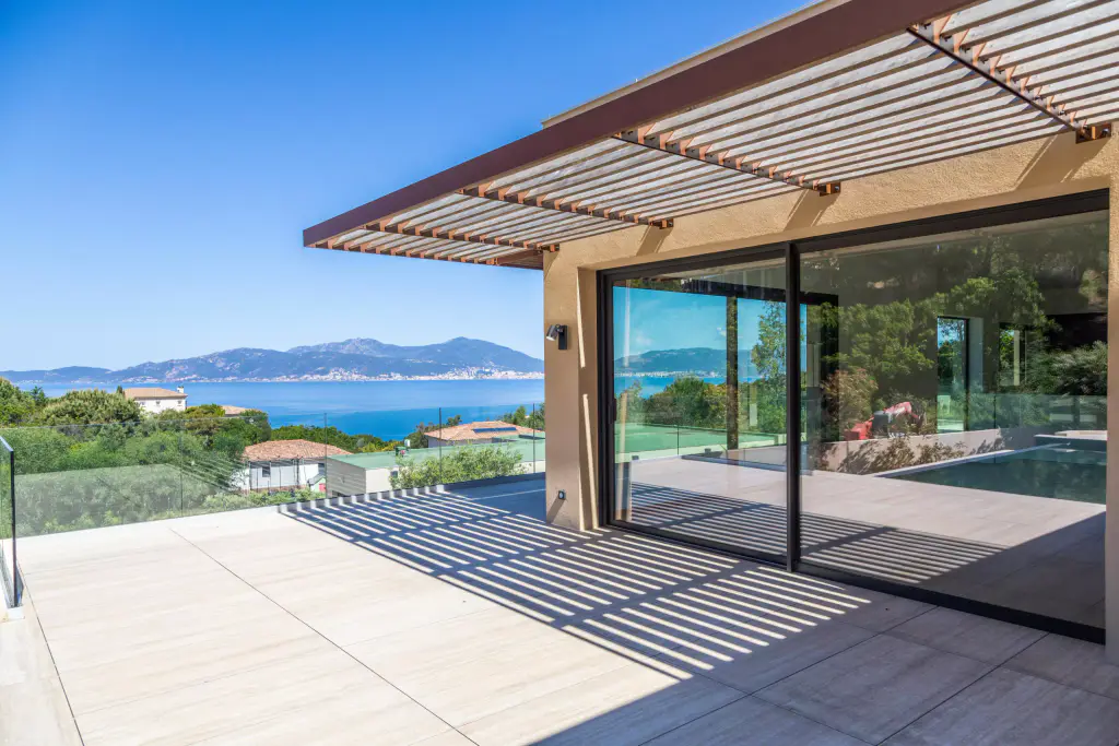 Brise-soleil en casquette à lames de bois sur consoles d’acier brun, au-dessus de la baie coulissante d’angle ; terrasse carrelée striée d’ombres, golfe et montagnes à gauche.
