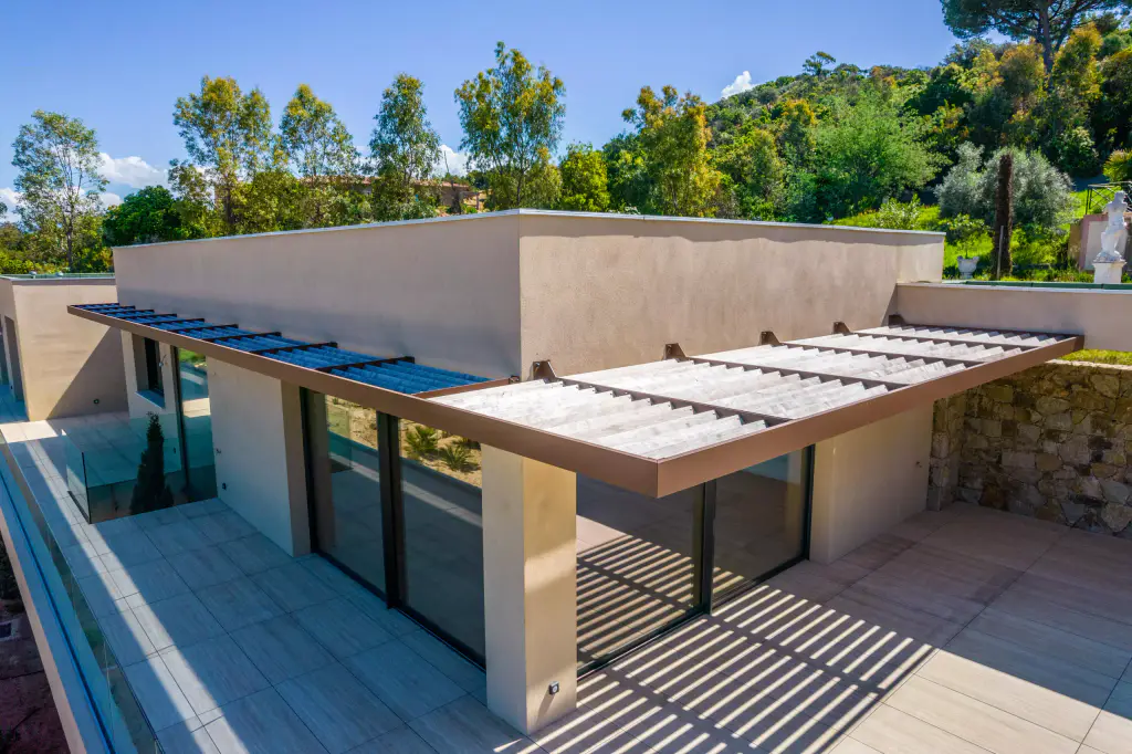 Vue drone de trois quarts : la casquette brise-soleil, lames de bois grisé et rive d’acier brun, longe deux façades de la villa ocre ; ombres rayées sur la terrasse carrelée.
