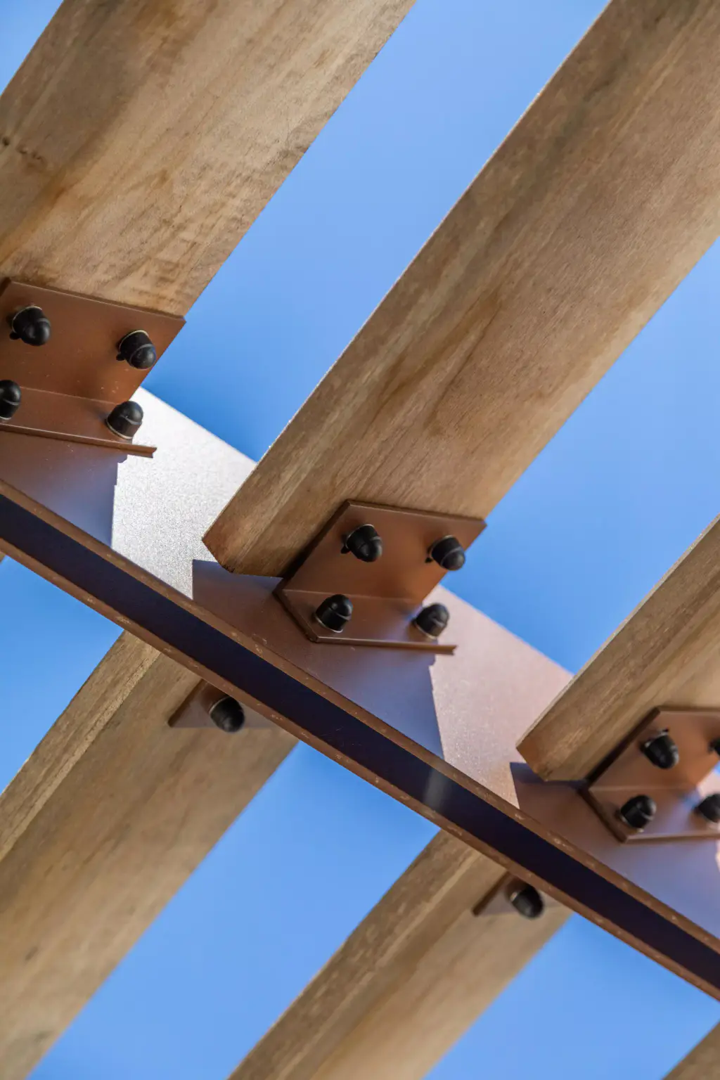 Très gros plan de la fixation du brise-soleil : équerres d’acier brun et boulons noirs tiennent les lames de bois sur la traverse, sur le ciel bleu.