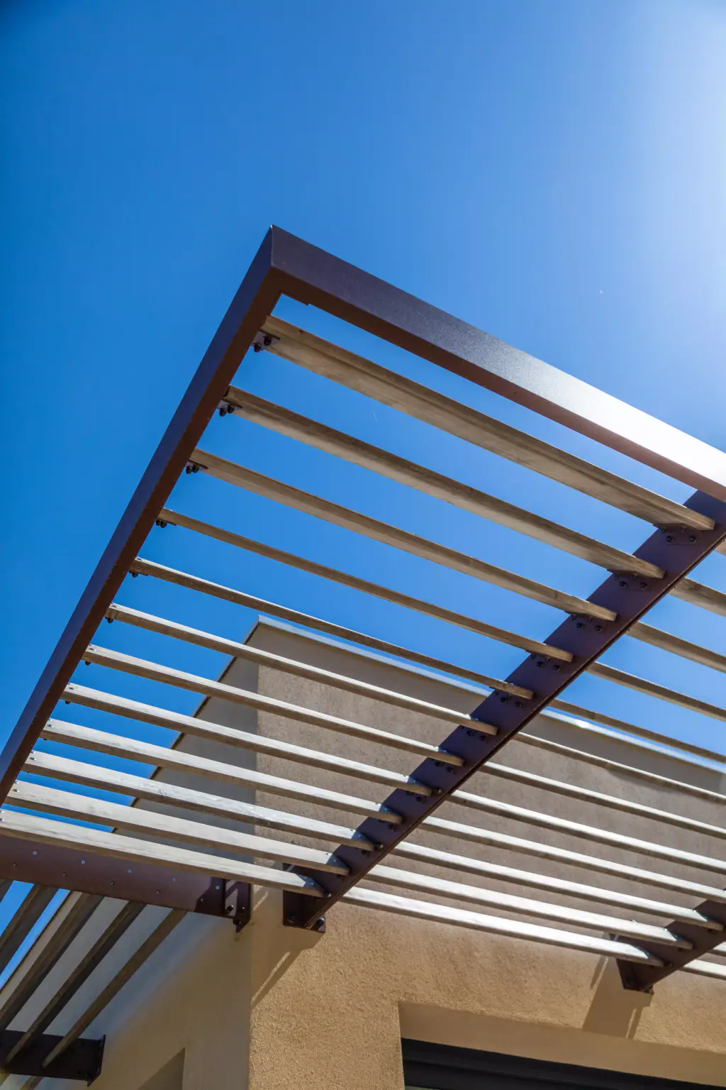 Angle du brise-soleil en contre-plongée : le cadre d’acier brun et les lames de bois clair se détachent en diagonale sur un ciel bleu profond, façade ocre en bas.
