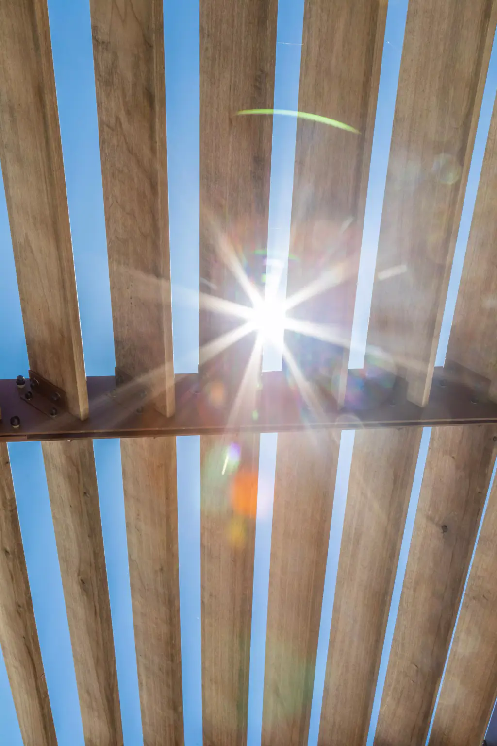 Lames de bois du brise-soleil vues de dessous : une étoile de soleil perce entre deux lames, ciel bleu entre les bandes.
