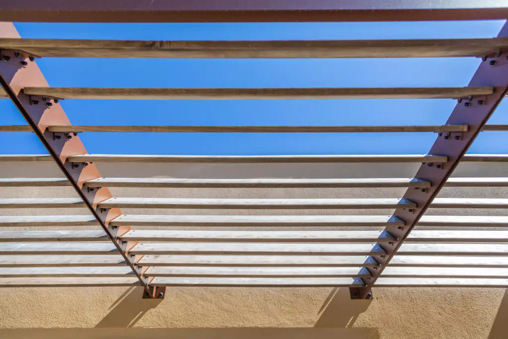 Brise-soleil vu de dessous, de face : les lames de bois horizontales filent entre deux consoles d’acier brun fixées à la façade ocre, le ciel bleu passe entre elles.