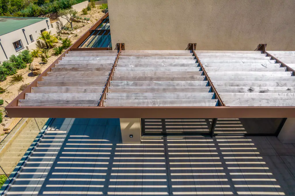 Vue drone plongeante : lames de bois grisé du brise-soleil et rive d’acier brun, tête de baie vitrée dessous, larges ombres rayées sur la terrasse, jardin sec à gauche.