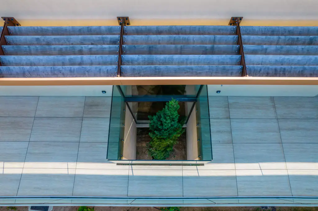 Vue drone plongeante : sous les lames de bois du brise-soleil et leurs consoles d’acier brun, un patio ceint de verre planté d’un cyprès s’ouvre dans la terrasse carrelée.