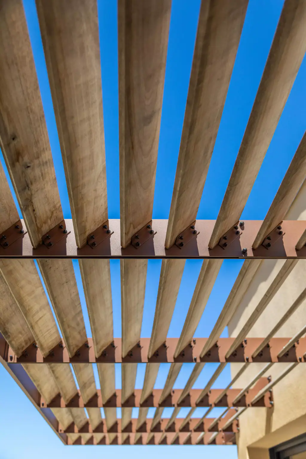 Sous le brise-soleil, en vertical : les lames de bois fuient vers le fond, trois rangs de platines d’acier brun boulonnées se répètent, angle de la façade ocre à droite.