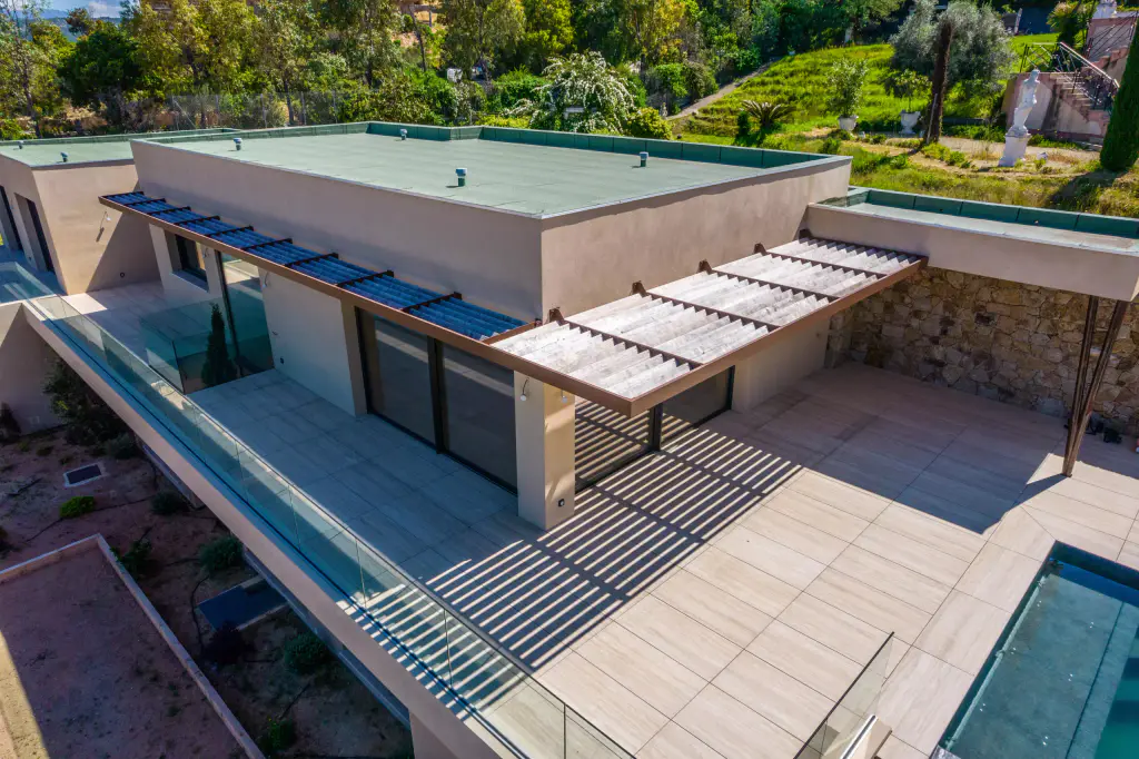 Vue drone de trois quarts en plongée : villa ocre à toit vert, casquette brise-soleil à lames de bois, terrasse carrelée rayée d’ombres, coursive vitrée, angle du bassin.