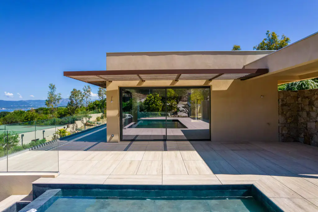 Villa ocre à toit plat vue depuis le bassin : brise-soleil en casquette à lames de bois au-dessus de la baie vitrée, terrasse carrelée, mur de pierre à droite, golfe à gauche.