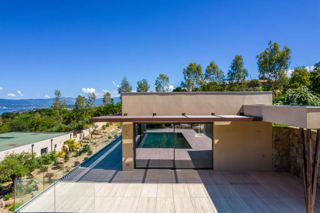 Vue drone basse de face : villa ocre à toit plat, casquette brise-soleil au-dessus de la baie où se reflète le bassin, terrasse rayée d’ombres, jardin sec et golfe à gauche.
