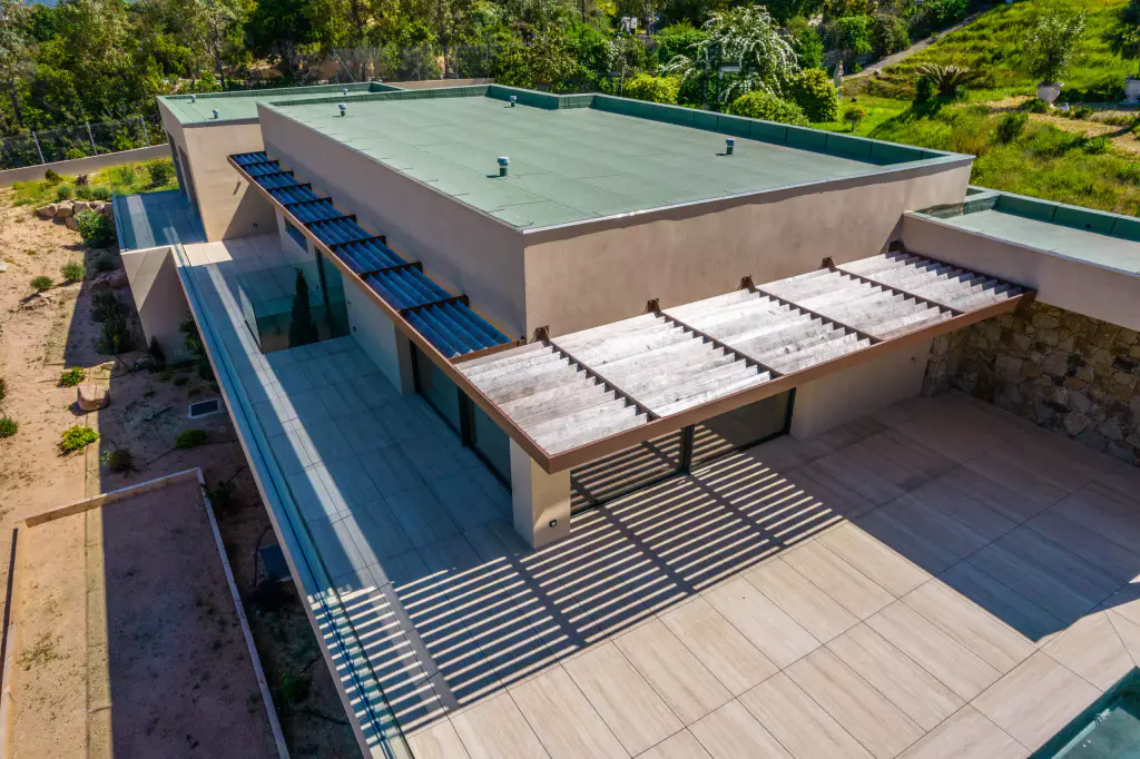 Vue drone de trois quarts : villa ocre à toit plat vert, casquette brise-soleil à lames de bois le long de deux façades, terrasse carrelée rayée d’ombres, mur de pierre, maquis.