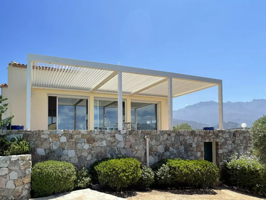 White pergola set against a pale yellow house, stone wall, clipped box, mountain.