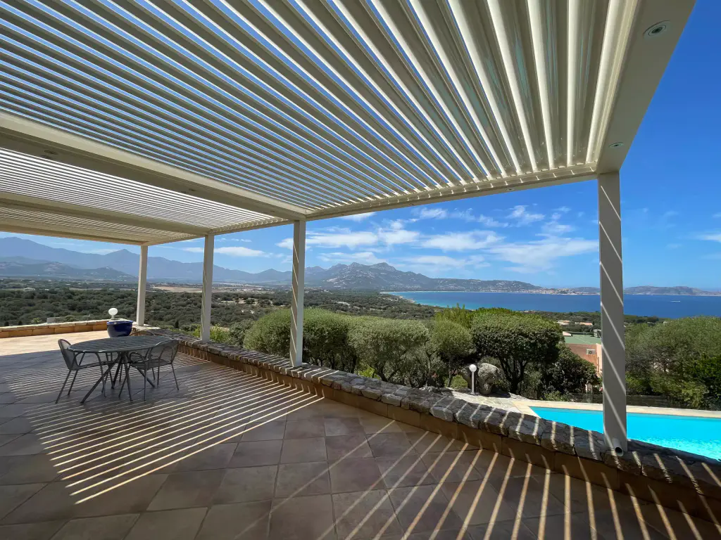 Sous la pergola blanche : lames entrouvertes, ombres rayées sur la terrasse en terre cuite, golfe et montagnes, table bistrot.