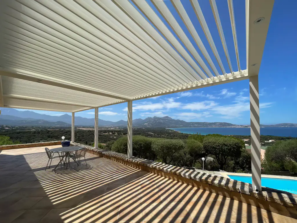 Pergola blanche à lames entrouvertes, ombres rayées, piscine en contrebas, golfe et montagnes.