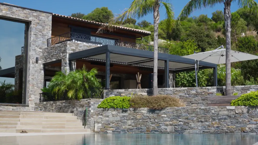 Film de la Casa Palma : les toits et la piscine vus du ciel, la villa de pierre et ses palmiers, le dessous de la pergola, les lames, leur ombre sur le parquet et la table, une main sur la télécommande, le store screen qui descend, puis les lames qui se ferment vues du ciel.