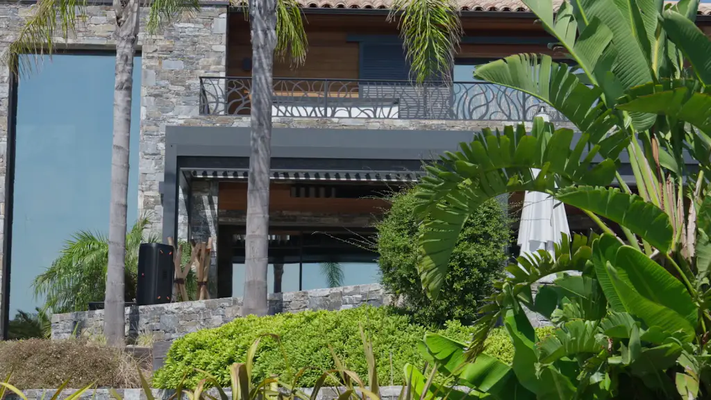 Film : une villa de pierre et de bois vue du jardin, des feuillages au premier plan, puis un store screen gris qui descend le long d’un poteau de la pergola.