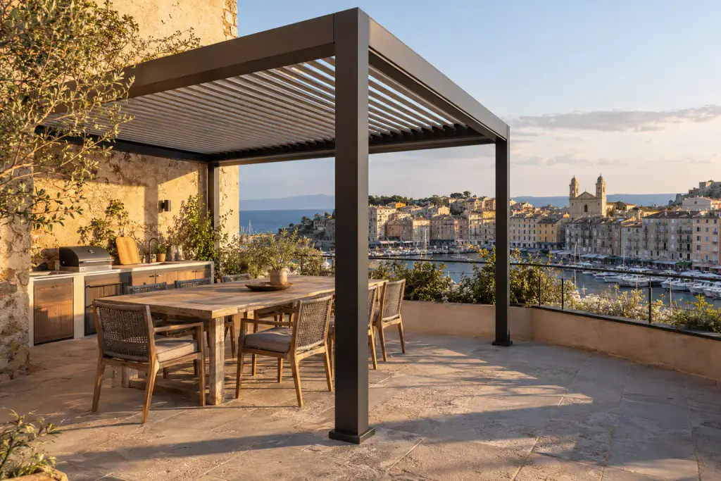 Pergola à lames en aluminium noir sur une terrasse dallée au-dessus d’un port, table en bois et chaises tressées, cuisine d’extérieur contre un mur de pierre, façades colorées et église à deux clochers au fond, lumière du soir.