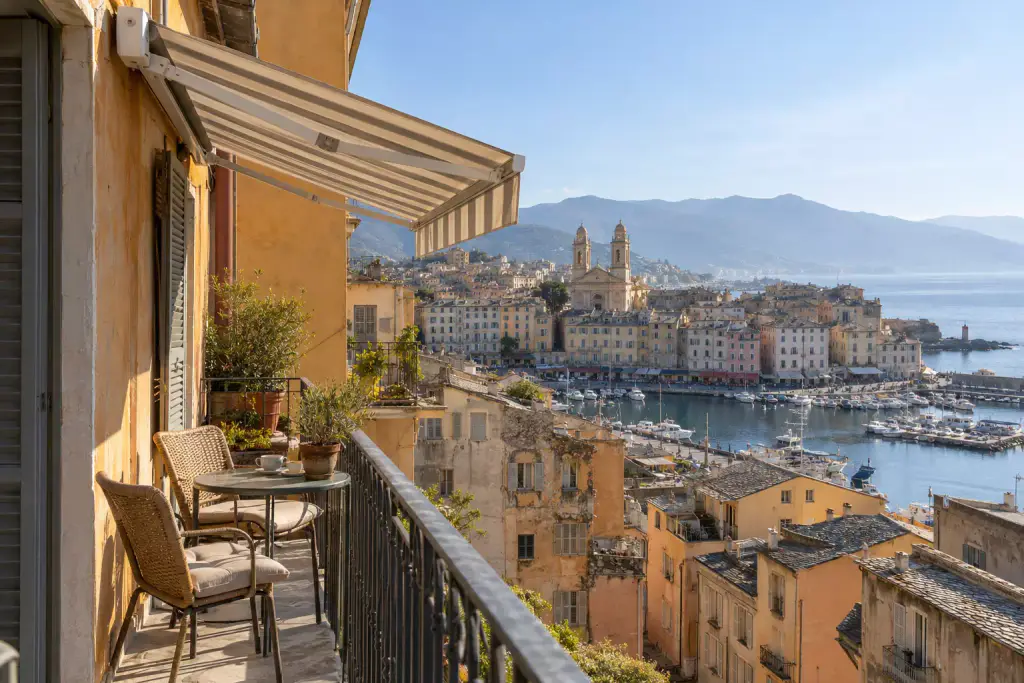 Balcon étroit d’un immeuble ocre sous un store banne à rayures écrues, petite table ronde et deux chaises en rotin, vue plongeante sur les toits, le port de plaisance et une église à deux clochers, montagnes au fond.