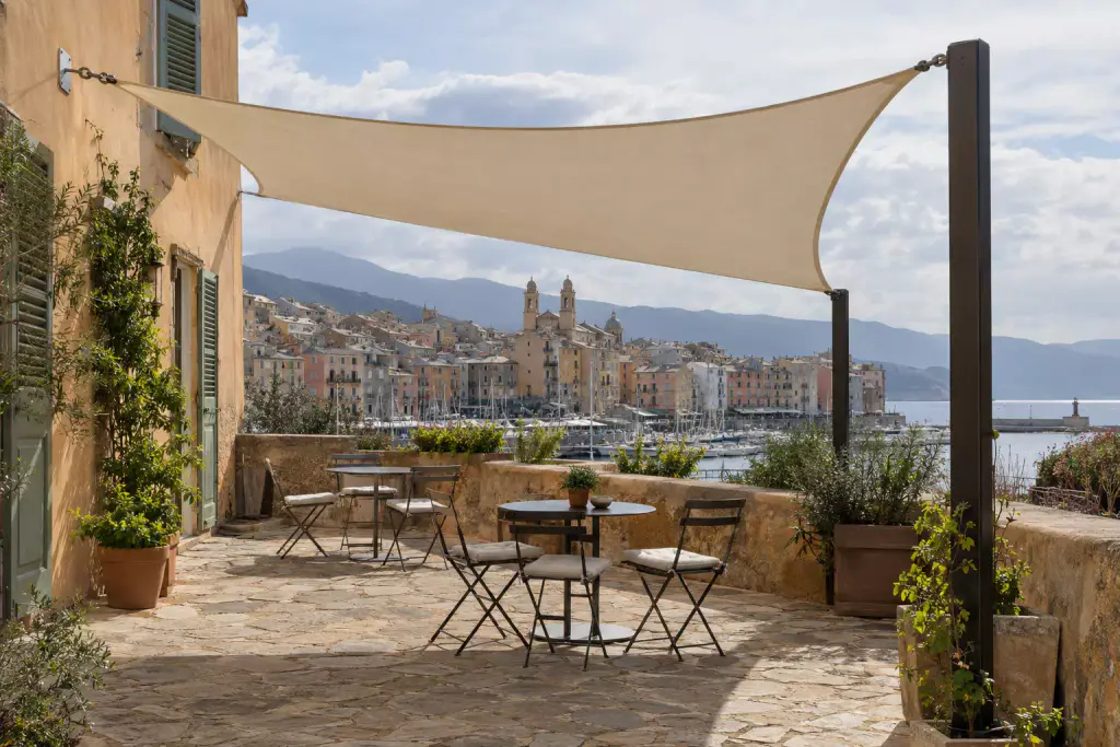 Voile d’ombrage beige tendue entre la façade ocre d’une maison et un mât métallique sombre, au-dessus d’une terrasse en pierre avec tables bistrot et chaises pliantes, port, façades colorées et église à deux clochers au fond.