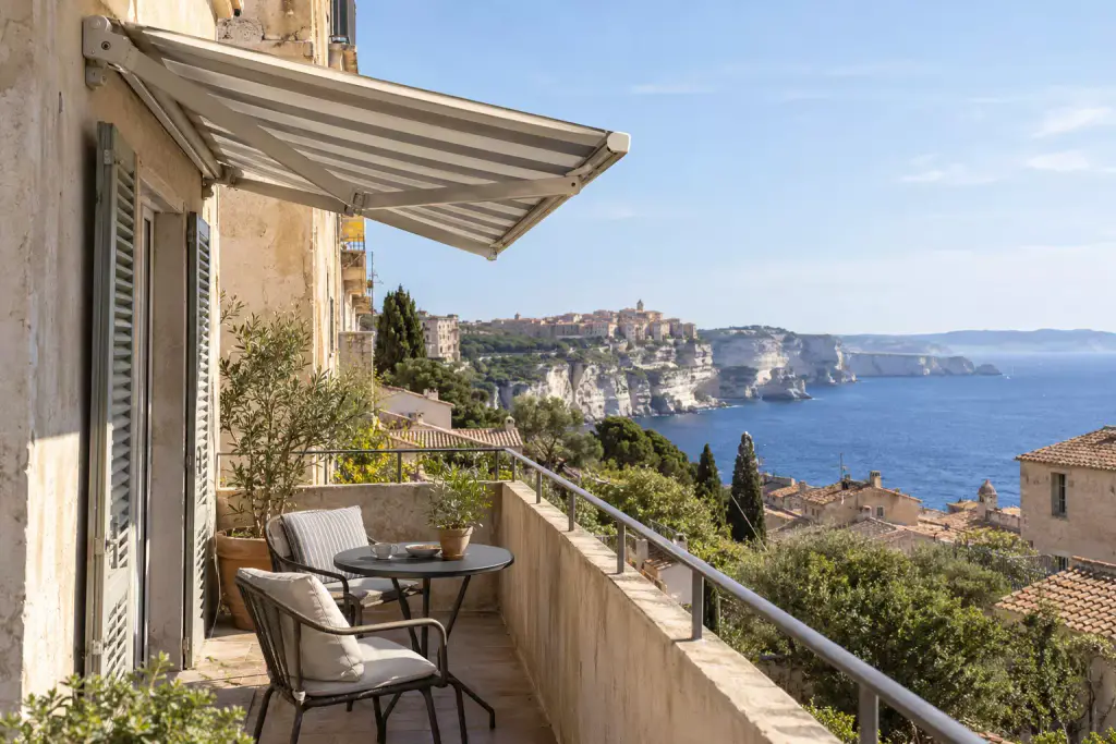 Store banne à rayures grises et blanches déroulé au-dessus d’un balcon étroit, avec un guéridon, deux chaises et des volets gris ; en contrebas, des toits de tuiles, et au loin la haute ville sur ses falaises blanches et la mer.