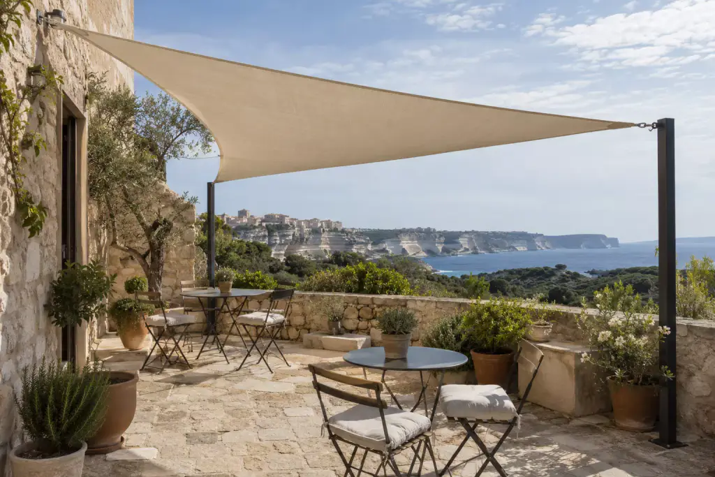Voile d’ombrage triangulaire beige tendue entre le mur de pierre d’une maison et deux mâts noirs, au-dessus d’une terrasse dallée avec des tables de bistrot et des pots ; au fond, les falaises blanches et la haute ville au-dessus de la mer.