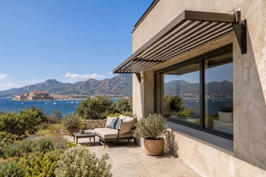 Brise-soleil à lames anthracite posé en casquette sur consoles au-dessus d’une grande baie vitrée, dans une façade enduite ; la vitre reflète les montagnes ; terrasse dallée, canapé et oliviers en pot ; à gauche, la citadelle de Calvi, la baie et des voiliers.