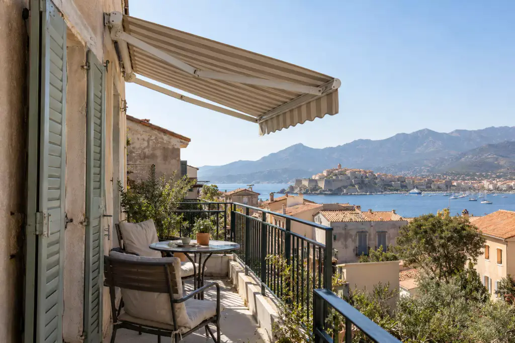 Store banne à rayures beige et blanc déployé au-dessus d’un balcon étroit, volets verts, petite table ronde et deux fauteuils ; en contrebas, des toits de tuiles, la baie, la citadelle de Calvi, la marina et les montagnes.