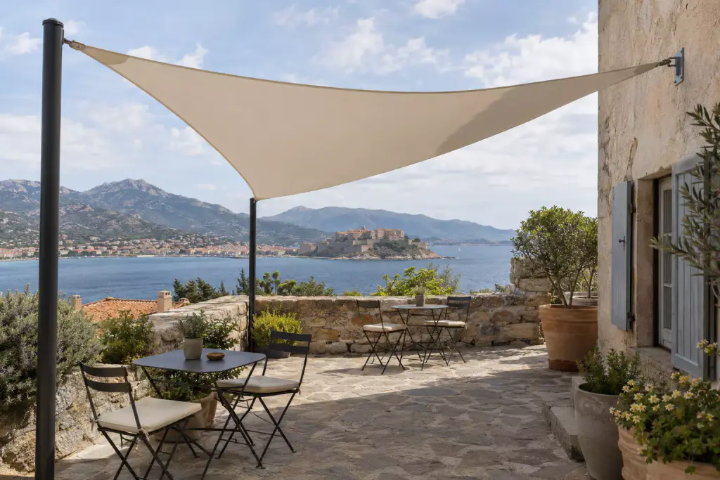 Voile d’ombrage triangulaire beige tendue entre deux mâts noirs et la façade en pierre d’une maison aux volets bleus ; terrasse dallée de pierre, tables et chaises pliantes, muret de pierre ; au fond, la citadelle de Calvi sur la baie et les montagnes.